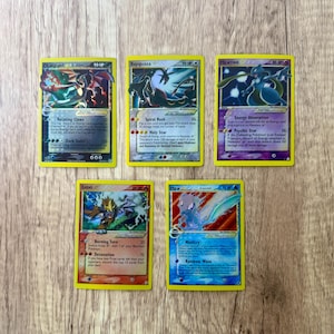 K&ouml;nnte beinhalten: F&uuml;nf Pok&eacute;mon-Sammelkarten mit gelben R&auml;ndern werden auf einer Holzoberfl&auml;che gezeigt. Die Karten zeigen verschiedene Pok&eacute;mon-Charaktere, darunter Glurak, Rayquaza, Mewtwo, Entei und Mew. Jede Karte hat einzigartige Kunstwerke und Text.