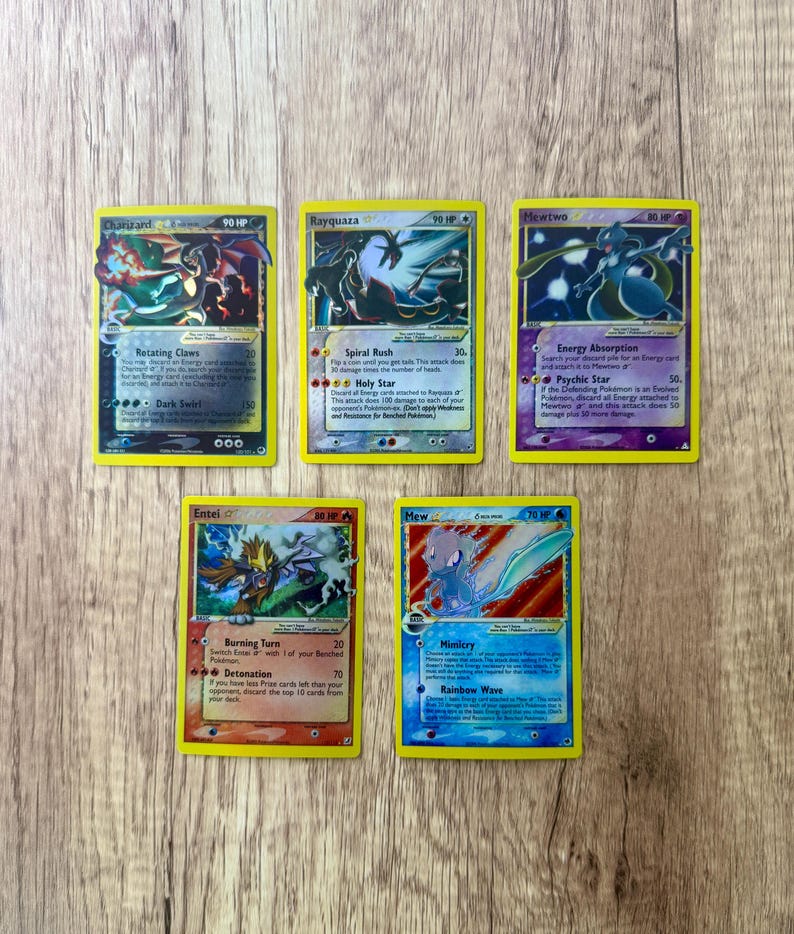 K&ouml;nnte beinhalten: F&uuml;nf Pok&eacute;mon-Sammelkarten mit gelben R&auml;ndern werden auf einer Holzoberfl&auml;che gezeigt. Die Karten zeigen verschiedene Pok&eacute;mon-Charaktere, darunter Glurak, Rayquaza, Mewtwo, Entei und Mew. Jede Karte hat einzigartige Kunstwerke und Text.