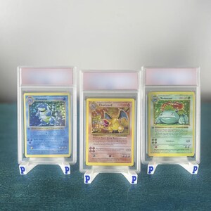 Peut inclure: Trois cartes à collectionner insérées dans des étuis de protection rectangulaires en plastique transparent, chacune posée sur un petit support blanc marqué d'un P bleu. Les cartes présentent des illustrations de Blastoise, Charizard et Venusaur.