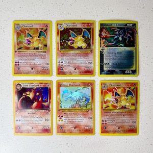 Peut inclure: Une collection de six cartes à collectionner Charizard disposées sur deux rangées. Les cartes présentent diverses illustrations de la créature orange ressemblant à un dragon, dont un Charizard brillant bleu, avec du texte imprimé détaillant les attaques, les PV et les statistiques.