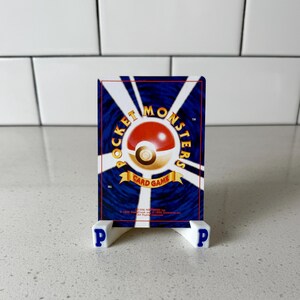 May include: A rectangular trading card featuring a dark blue background with white radiating lines and a central red and white sphere. The text reads POCKET MONSTERS CARD GAME, 1996 Nintendo, 1996 Game Freak inc, 1996 Creatures inc. It rests on a white stand.