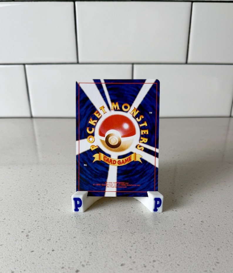 May include: A rectangular trading card featuring a dark blue background with white radiating lines and a central red and white sphere. The text reads POCKET MONSTERS CARD GAME, 1996 Nintendo, 1996 Game Freak inc, 1996 Creatures inc. It rests on a white stand.