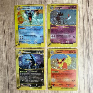 Może przedstawiać: Cztery karty kolekcjonerskie Pokémon z Gyarados, Gengar, Umbreon i Flareon. Każda karta ma unikalną ilustrację Pokémona, z tekstem opisującym jego umiejętności i HP. Karty leżą na drewnianej powierzchni.