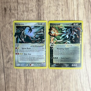 Op de afbeelding: Twee Pokémon-verzamelkaarten met Rayquaza en Charizard. De Rayquaza-kaart heeft een zwart-blauwe illustratie met tekst. De Charizard-kaart heeft een vurige illustratie met tekst. Beide kaarten hebben gele randen.