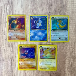 Peut inclure: Cinq cartes à collectionner Pokémon vintage, dont Dark Charizard, Dark Blastoise, Dark Dragonite, Dark Machamp et Dark Raichu. Chaque carte a une bordure jaune et des illustrations uniques de Pokémon. Les cartes sont posées sur une surface en bois.