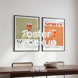 Könnte beinhalten: Zwei gerahmte Poster mit Cocktail-Illustrationen hängen an einer weißen Wand. Ein Poster zeigt ein Getränk mit einer Orangenscheibe, das andere ein ähnliches Getränk. Die Poster befinden sich über einem dunklen Holztisch mit einer Schüssel und Stühlen.