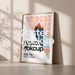 Op de afbeelding: Een ingelijste poster-mockup met een kleurrijke illustratie. Het kunstwerk toont een gebouw met een oranje dak, een brug en een geometrisch ontwerp in roze en wit. De tekst "Poster Mockup" en "PSD File Din A Ratio" zijn zichtbaar.