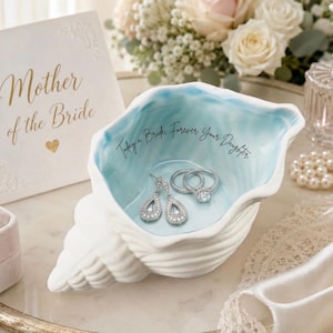 May include: A white seashell-shaped jewelry dish with a light blue interior, holding a pair of drop earrings and two rings. The dish has the inscription "Today a Bride, Forever Your Daughter." A card that says "Mother of the Bride" is also present.