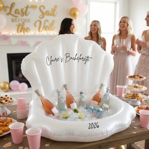 May include: An inflatable white seashell-shaped beverage cooler filled with ice, bottles of rosé wine, and water bottles. The cooler is on a wooden table with pink cups and snacks. The text "Claire's Bachelorette" and "2026" are visible.