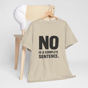May include: A beige t-shirt with the words "NO IS A COMPLETE SENTENCE" in black block letters. The shirt hangs on a wooden hanger. A white baseball cap rests on a wooden chair.