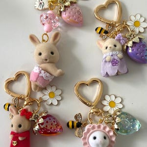 May include: A collection of whimsical keychains featuring miniature animal figures, including bunnies and cats, adorned with strawberry and flower charms. Each keychain has a gold-colored heart-shaped clasp and decorative bee accents.