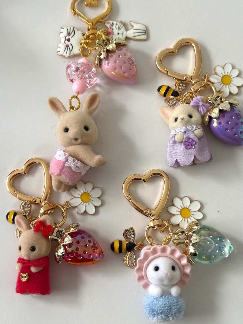 May include: A collection of whimsical keychains featuring miniature animal figures, including bunnies and cats, adorned with strawberry and flower charms. Each keychain has a gold-colored heart-shaped clasp and decorative bee accents.