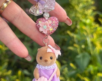 Llavero Sylvanian de nutria hecho a mano: vestido morado, lazo rosa, cuentas decorativas.