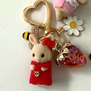 May include: A gold-colored heart-shaped keychain with a bunny charm wearing a red dress and bow. The keychain also features a bee, a strawberry, and a daisy charm. The bunny is beige with black eyes.