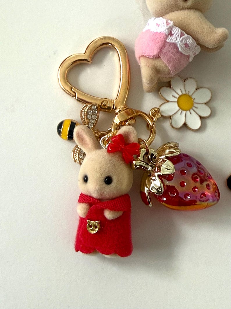 May include: A gold-colored heart-shaped keychain with a bunny charm wearing a red dress and bow. The keychain also features a bee, a strawberry, and a daisy charm. The bunny is beige with black eyes.
