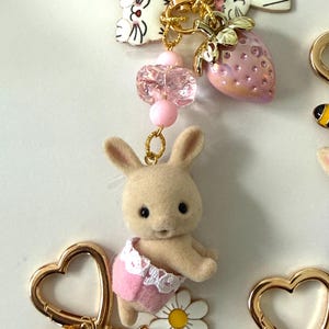 May include: A collection of whimsical keychains featuring a variety of charms. The keychains include a small bunny wearing a pink diaper, a strawberry, a daisy, and a bee. The keychains are attached to gold-colored heart-shaped clasps.