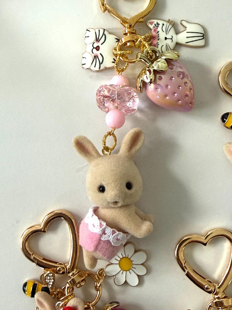 May include: A collection of whimsical keychains featuring a variety of charms. The keychains include a small bunny wearing a pink diaper, a strawberry, a daisy, and a bee. The keychains are attached to gold-colored heart-shaped clasps.