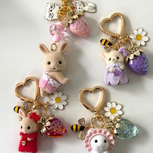 May include: A collection of whimsical keychains featuring miniature animal figures, including bunnies and a cat, adorned with charms like strawberries, flowers, and bees. Each keychain has a gold heart-shaped clasp.