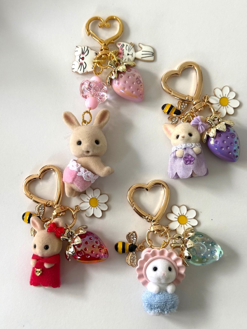 May include: A collection of whimsical keychains featuring miniature animal figures, including bunnies and a cat, adorned with charms like strawberries, flowers, and bees. Each keychain has a gold heart-shaped clasp.
