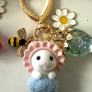 May include: A gold-colored heart-shaped keychain with a white bunny charm wearing a pink bonnet and blue dress. The keychain also features a bee charm, a daisy charm, and a clear strawberry charm.