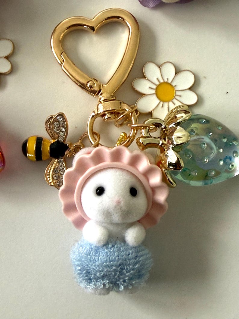 May include: A gold-colored heart-shaped keychain with a white bunny charm wearing a pink bonnet and blue dress. The keychain also features a bee charm, a daisy charm, and a clear strawberry charm.