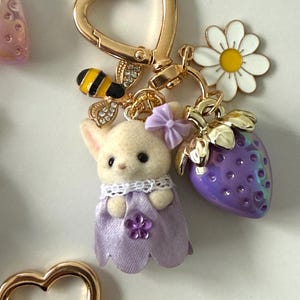 May include: A gold-colored heart-shaped keychain with a small bunny wearing a purple dress, a bee, a white daisy, and a purple strawberry charm. The bunny has a purple bow and a flower on its dress.