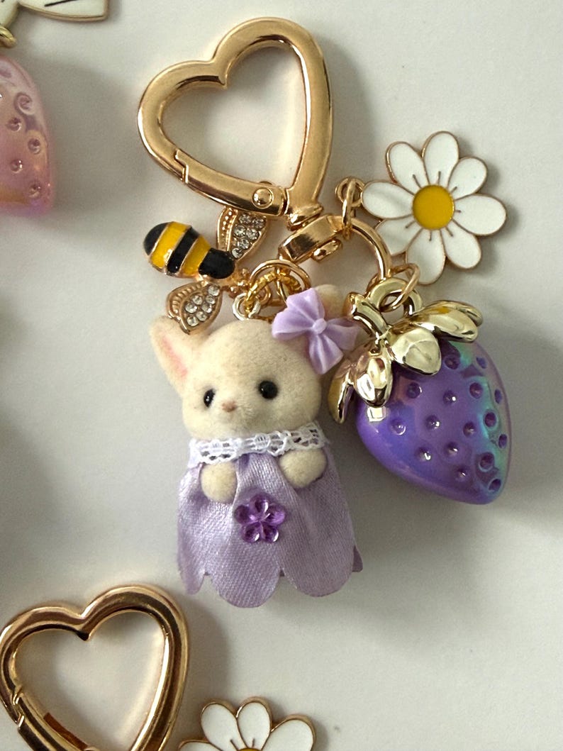 May include: A gold-colored heart-shaped keychain with a small bunny wearing a purple dress, a bee, a white daisy, and a purple strawberry charm. The bunny has a purple bow and a flower on its dress.