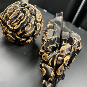 May include: A spherical articulated object and an elongated, segmented, flexible object, both featuring a black base color with textured, raised golden-brown patterns resembling snake scales or a reptilian skin texture throughout their surfaces.