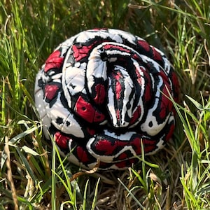 May include: A coiled snake figurine featuring a high-contrast pattern of vibrant red, stark white, and deep black scales. The sculpture is rendered with a textured, segmented surface that mimics the appearance of a resting reptile in a tight circle.