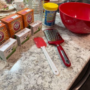 May include: A selection of cleaning and kitchen items on a work surface. Includes boxes of baking soda, Dove soap bars, a red mixing bowl, a grater, a spatula, and a container of Oxiclean.