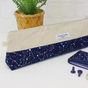 Może przedstawiać: Etui na igły wykonane z niebieskiego i białego materiału z wzorem konstelacji i tekstem "Coralie Green Designs". Etui jest otwarte i zawiera dwa małe fioletowe uchwyty na igły oraz srebrne nożyczki.
