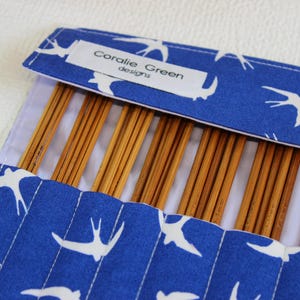 May include: A blue fabric knitting needle case with a white bird pattern, holding several wooden knitting needles. The label reads "Coralie Green designs". The case is open, revealing the needles.