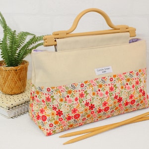 Puede incluir: Bolso tote color crema con un estampado floral en la mitad inferior y un asa de madera. La bolsa tiene una etiqueta que dice "Carole Green Studio". Dos agujas de tejer de madera descansan frente a la bolsa.