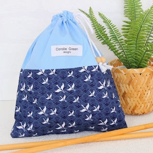May include: A blue and navy drawstring bag with a white crane pattern. The top is light blue, and the bottom features a navy wave pattern with white cranes. The bag has a white drawstring and a tag that reads "Coralie Green designs."
