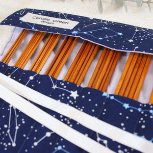 Puede incluir: Un estuche azul marino para agujas de tejer con un patrón de constelaciones. El estuche está abierto, revelando varias agujas de tejer de madera. Cintas blancas aseguran el estuche. La etiqueta dice "Coralie Green designs."