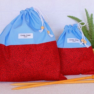 Könnte beinhalten: Zwei Projektbeutel mit Kordelzug in Blau und Rot mit schwarzen Tupfen und zwei Stricknadeln aus Holz. Die Taschen haben weiße Kordelzüge mit Holzperlen und ein Etikett mit der Aufschrift "Corolie Green designs."