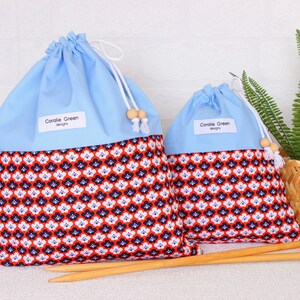 May include: Two drawstring project bags with a light blue top and a patterned bottom in red, white, and navy blue. The bags have white drawstrings with wooden beads. The text "Coralie Green designs" is on a white label on each bag. Two wooden knitting needles are in front.