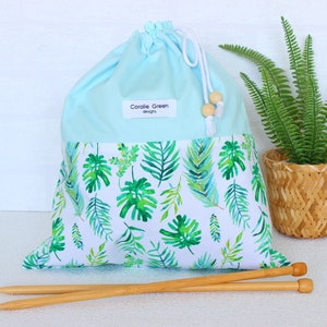 May include: A light blue drawstring project bag featuring a white lower section with a green tropical leaf pattern. A white label reads Coralie Green designs. The bag has white cords with wooden beads and is paired with two wooden knitting needles.