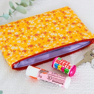 Op de afbeelding: Een gele etui met bloemenprint en een rode rits, naast een lippenbalsem tube, een rol snoep en een sleutel met een label "Rhythm is a Dancer".