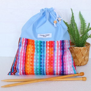 Op de afbeelding: Een lichtblauwe tas met trekkoord en een levendig, meerkleurig verticaal streeppatroon met witte stippen op de onderste helft. Een wit label met de tekst Coralie Green designs is in het midden geplaatst. Twee houten breinaalden liggen ervoor.