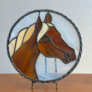May include: Stained glass artwork depicting a horse's head in shades of brown, tan, and white, set within a circular frame. The horse's profile is detailed, with a textured mane and a dark, decorative border. The artwork is displayed on a small metal stand.