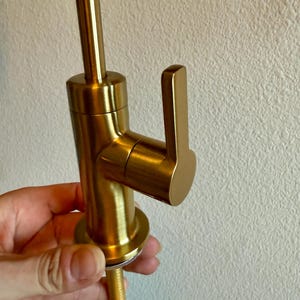 May include: A close-up of a gold-colored faucet with a lever handle. The faucet has a cylindrical body and a long, threaded base. The handle is rectangular and angled. The faucet is held by a person's hand.