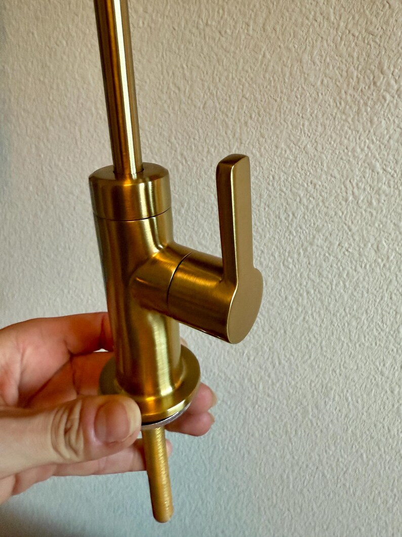 May include: A close-up of a gold-colored faucet with a lever handle. The faucet has a cylindrical body and a long, threaded base. The handle is rectangular and angled. The faucet is held by a person's hand.