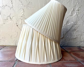 May include: Two off-white lamp shades with a pleated fabric design. The top shade is smaller and rests at an angle on top of the larger, wider base shade. Both shades feature a consistent vertical pleat pattern and a smooth, finished edge.