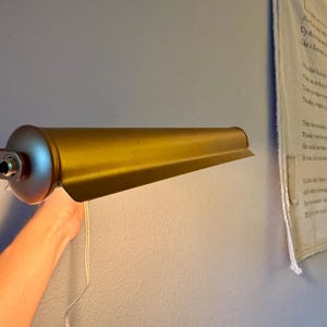 May include: A gold-colored, vintage-style wall-mounted lamp. The lamp has a cylindrical base with a black on/off switch and a long, tapered shade. A white cord hangs from the lamp. The lamp is mounted on a light blue wall.