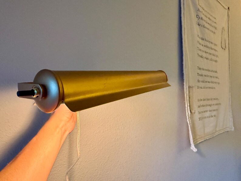 May include: A gold-colored, vintage-style wall-mounted lamp. The lamp has a cylindrical base with a black on/off switch and a long, tapered shade. A white cord hangs from the lamp. The lamp is mounted on a light blue wall.