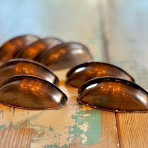 May include: A collection of antique-style, bronze-colored cup pulls. The pulls are arranged on a distressed wooden surface, showcasing their curved design and metallic sheen. These hardware pieces are ideal for cabinets and drawers.