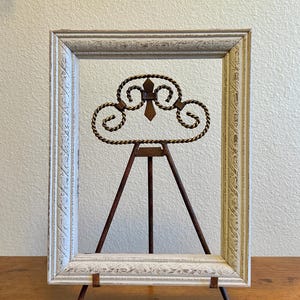 May include: A white rectangular picture frame with ornate detailing and a distressed finish. A decorative metal accent in the center features a fleur-de-lis design. The frame is supported by a small, rusty-brown metal easel.