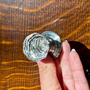 May include: A clear, faceted glass doorknob with a polished metal base. The knob is held in a hand, showcasing its intricate design and reflective qualities. The background is a wooden surface.