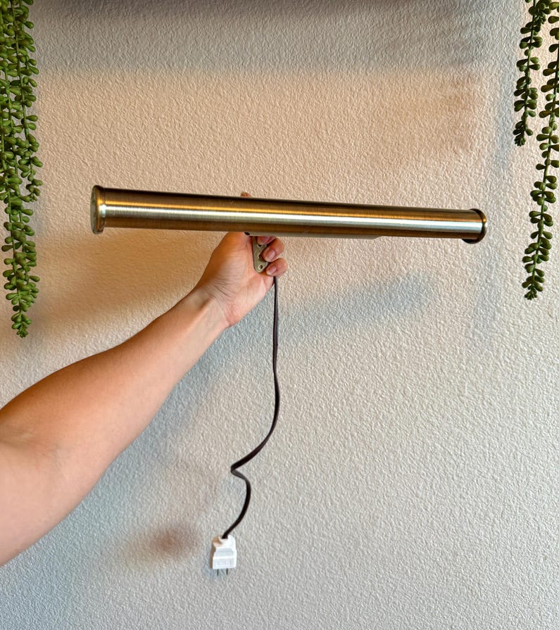 May include: A brass-colored, cylindrical wall-mounted light fixture with a black power cord and a white plug. The fixture is held by a hand against a textured, off-white wall, with hanging greenery on either side.